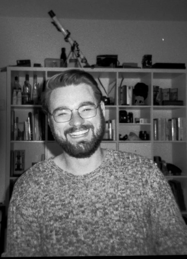 A B&W portrait of a man, bearded haphazardly smiling in front of a weird shelf